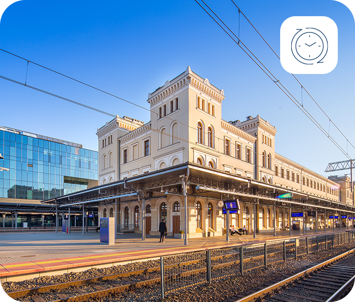 Dworzec kolejowy z peronami i torami w słoneczny dzień, w rogu ikona zegara symbolizująca punktualne podróże.